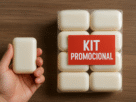 Mão segurando um sabonete ao lado de um kit promocional com seis unidades embaladas e etiqueta vermelha “KIT PROMOCIONAL” sobre mesa de madeira.