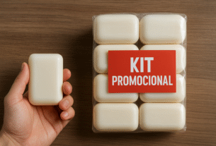 Mão segurando um sabonete ao lado de um kit promocional com seis unidades embaladas e etiqueta vermelha “KIT PROMOCIONAL” sobre mesa de madeira.